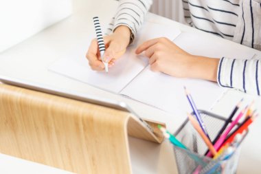 Not defterine kalemle ödev yazan çocuk ellerini kapat. Evde dijital tablet bilgisayarla masada oturan çocuk