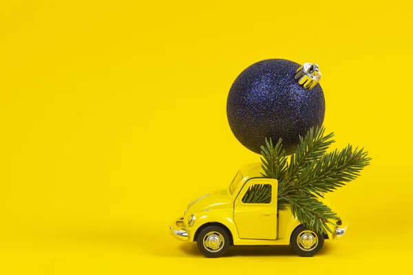 Christmas background. Little retro toy model car with small Christmas tree branch and navy decoration bauble ball on yellow background