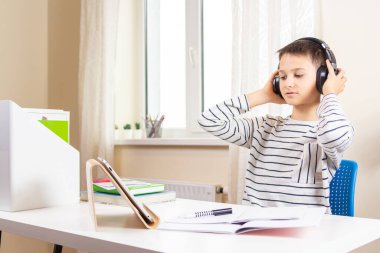 Tablet bilgisayarı olan bir çocuk elinde kitaplarla masada oturuyor ve video görüşmesi yapıyor, evde sanal kiralama var.