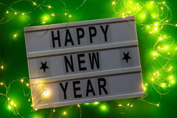Белый лайтбокс с поздравлением с Новым годом и гирляндой на зеленом фоне. Вид сверху — стоковое фото