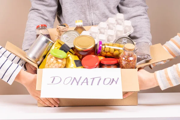 Voluntarios manos sosteniendo caja de donaciones de alimentos con productos de comestibles en escritorio blanco — Foto de Stock