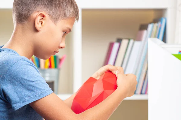 Мальчик смотрит видеоуроки и делает красное полигональное бумажное сердце. Образование, обучение, бумажные ремесла, развлечения дома — стоковое фото