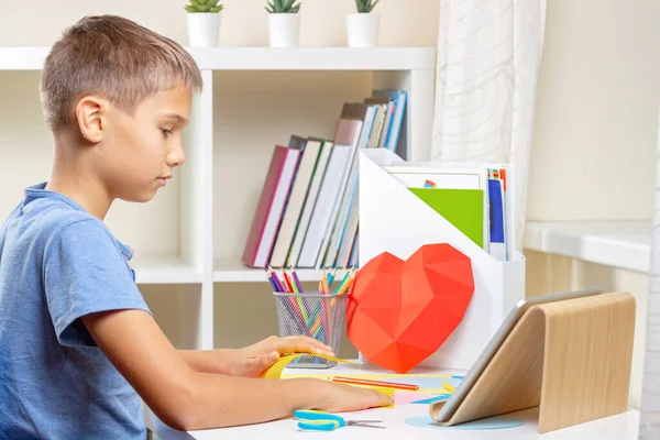Menino assistindo vídeo tutorial e fazendo vermelho papel poligonal coração. Educação, aprendizagem, artesanato de papel, entretenimento em casa — Fotografia de Stock