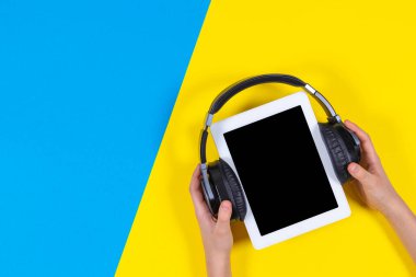 Çevrimiçi öğrenme, uzaktan eğitim, e-öğrenme konsepti. Çocuk elleri mavi ve sarı arka planda kablosuz kulaklıklı dijital tableti tutuyor. Üst görünüm