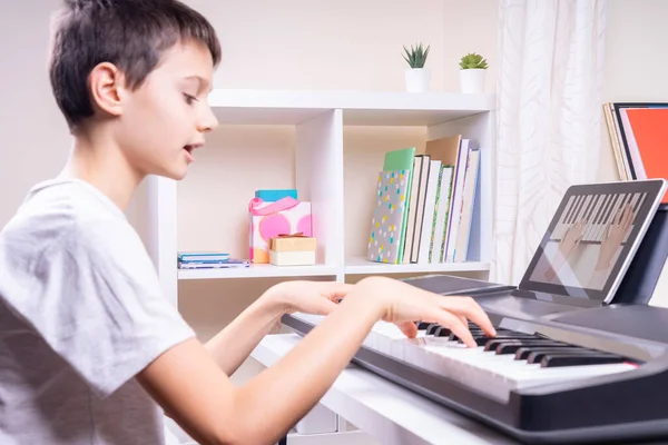 Online learning, remote education. Boy watching video tutorial at tablet computer, playing digital piano and singing at home