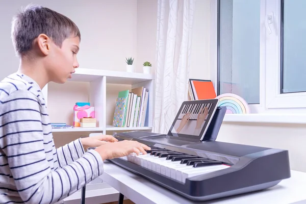 Online learning, remote education. Boy watching video lesson at digital tablet computer, playing digital piano and singing at home