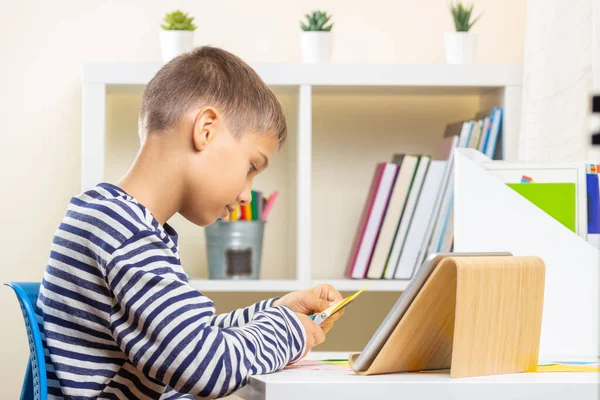 ハサミで色のついた紙を切る少年。子供はビデオチュートリアルを見て、自宅で紙工芸プロジェクトを作る — ストック写真