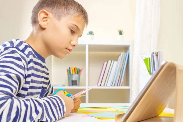 Educación, aprendizaje, artesanía del papel, entretenimiento. Chico de corte de papel de color con tijeras y mirando video tutorial en la computadora de la tableta en casa — Foto de Stock