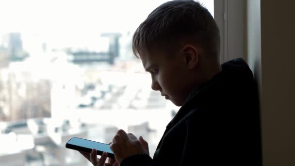 Θλιβερός έφηβος με μαύρο μπουφάν κάθεται στο περβάζι του παραθύρου, κοιτάζοντας έξω από το παράθυρο και χρησιμοποιώντας το κινητό τηλέφωνο για τα μέσα κοινωνικής δικτύωσης, παρακολουθήστε βίντεο. Κοινωνική απόσταση κατά τη διάρκεια πανδημίας αποκλεισμού στο σπίτι — Αρχείο Βίντεο