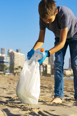 Plastik çöpten şehir plajını temizliyorum. Çocuk plajdan plastik şişe çöpü topluyor ve geri dönüşüm için plastik torbaya koyuyor.