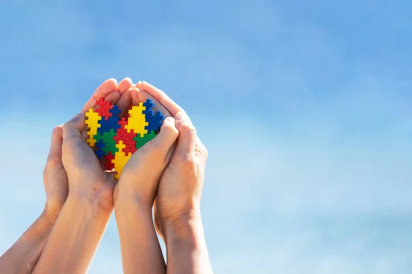Autista gyerek kezek sokszínű puzzle szív által támogatott anya kezét kék ég háttér. Az autizmus világnapja — Stock Fotó