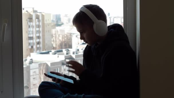 Adolescente con auriculares blancos en chaqueta negra con capucha sentado en el alféizar de la ventana y disfrutar escuchando música desde su teléfono celular. Distanciamiento social, entretenimiento en casa — Vídeo de stock