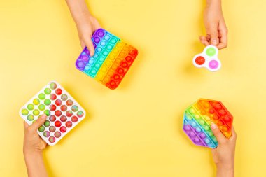 Bir çok el sarı arka planda hareketli oyuncaklar tutuyor. Pop-it kıpır kıpır oyunu stresi, endişeyi, otizmi azaltır, çocuklara duyusal ve dokunsal deneyim sağlar