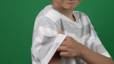 Genç bir çocuk yeşil ekran arka planında aşıdan sonra alçıyla koluna gösteriyor. Çocuklar ve gençler için aşı ve sağlık hizmetleri