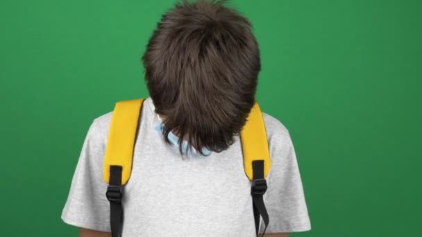 Adolescente con máscara de protección médica y mochila bajó la cabeza y miró hacia abajo. Niño de pie cerca de pantalla verde croma clave de fondo. Vídeo 4k — Vídeos de Stock