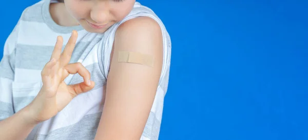 Genç çocuk, kolundaki bandaj bandını göstermiş. Mavi arka plandaki aşıdan sonra iyi görünüyor. Enjeksiyon covid aşı, çocuklar için sağlık hizmeti.