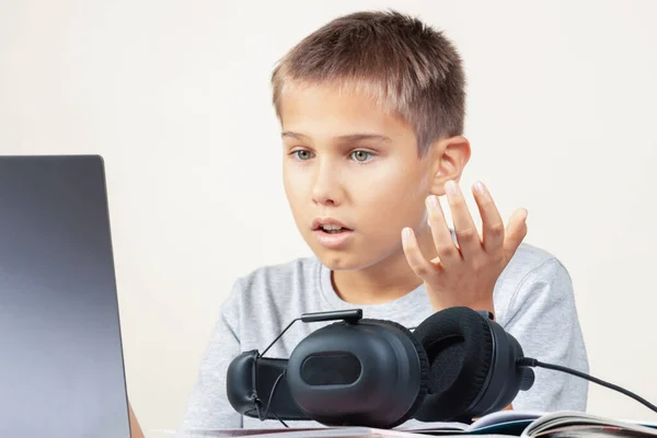 Criança chateada irritada chocada com computador portátil e fones de ouvido sentados à mesa com livros. Educação, aprendizagem online, jogos, comunicações de vídeo em casa — Fotografia de Stock