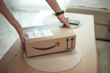 Alicante, İspanya - 02 Ocak 2026: Kadın elleriyle Amazon ambalaj kutusunu evdeki sehpanın üzerine koyuyor.