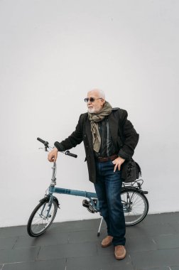 Beyaz bir duvarın yanında bisikletle duran yaşlı, gri saçlı hippi görünümlü bir iş adamı. İşe git, mola ver. Yaşlılar yaşam konsepti.