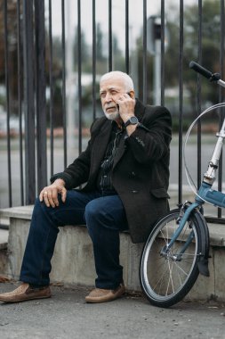 Kıdemli, kır saçlı iş adamı yanında bisikletle parapette otururken akıllı telefon uygulamasını kullanıyor. Hippi işadamı işe gidiyor. Yüze odaklan.