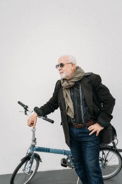 Beyaz bir duvarın yanında bisikletle duran yaşlı, gri saçlı hippi görünümlü bir iş adamı. İşe git, mola ver. Yaşlılar yaşam konsepti.