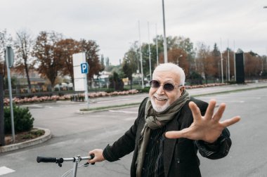 Hipster görünümlü yaşlı, gri saçlı bir iş adamı bisiklete binip kameraya el sallıyor. İşe git, mola ver. Yaşlılar yaşam konsepti. Yüze odaklan.