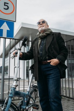Beyaz bir duvarın yanında bisikletle duran yaşlı, gri saçlı hippi görünümlü bir iş adamı. İşe git, mola ver. Yaşlılar yaşam konsepti.