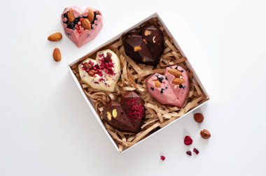 El yapımı çikolatalar, pembe, süt, siyah ve beyaz çikolatalı, çilekli, yabanmersinli ve bademli, mukavva bir kutuda, üst görünümde, beyaz bir arka planda.