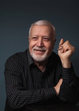 Yaşlı yakışıklı karizmatik adam elinde gözlüklerle oturuyor ve gülümsüyor, stüdyo, karanlık arka plan, portre