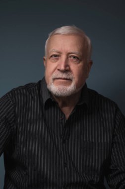 Yaşlı yakışıklı karizmatik adam elinde gözlüklerle oturuyor ve gülümsüyor, stüdyo, karanlık arka plan, portre