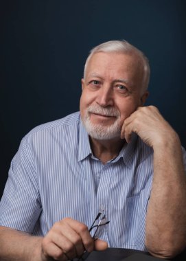 Yaşlı yakışıklı karizmatik adam elinde gözlüklerle oturuyor ve gülümsüyor, stüdyo, karanlık arka plan, portre