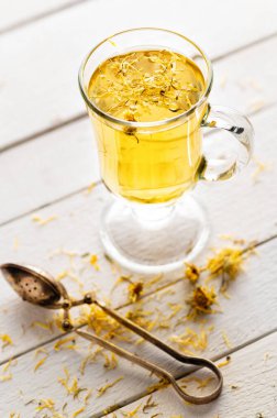 Kurumuş sarı çiçekleri ve süzgeci olan beyaz ahşap arka planda bir bardak aromatik çiçek çayı, yatıştırıcı, ilaç, antipiretik.