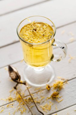 Kurumuş sarı çiçekleri ve süzgeci olan beyaz ahşap arka planda bir bardak aromatik çiçek çayı, yatıştırıcı, ilaç, antipiretik.
