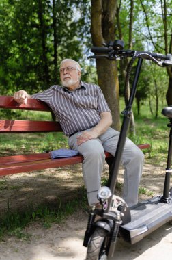 Olumlu, yaşlı bir adam parkta yürüyüşe çıktı, durdu ve elektrikli bir scooterın yanındaki bankta dinlenmek için oturdu. Yaz, güneşli bir gün. Yaşlıların sağlıklı yaşam tarzı.