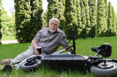 Olumlu, yaşlı bir adam parkta yürüyüşe çıktı, durdu ve elektrikli bir scooterın yanındaki çimlerin üzerinde dinlenmek için uzandı. Yaz, güneşli bir gün. Yaşlıların sağlıklı yaşam tarzı.