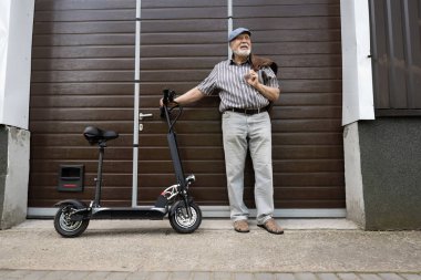 Olumlu, yaşlı bir adam kapının yanında duruyor ve direksiyonun arkasında bir eko scooter tutuyor. Yaşlıların sağlıklı yaşam tarzı.