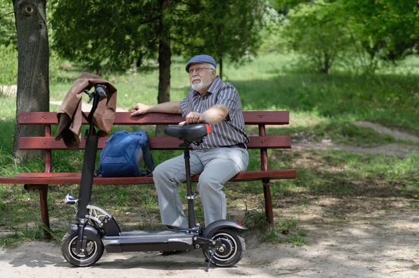 A positive, elderly man went for a walk to the park, stopped and sat down to rest on a bench next to an electric scooter. Summer, sunny day. Healthy lifestyle of the elderly.