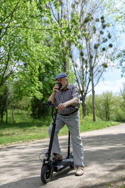 A positive, charismatic elderly man on an eco scooter went for a walk in the park. Drinks a drink from an eco paper cup. Healthy lifestyle of the elderly.