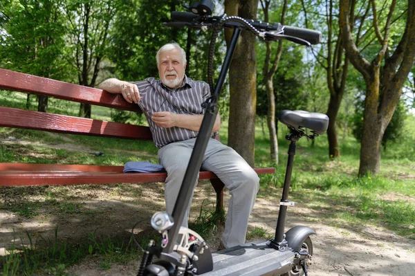 Olumlu, yaşlı bir adam parkta yürüyüşe çıktı, durdu ve elektrikli bir scooterın yanındaki bankta dinlenmek için oturdu. Yaz, güneşli bir gün. Yaşlıların sağlıklı yaşam tarzı.