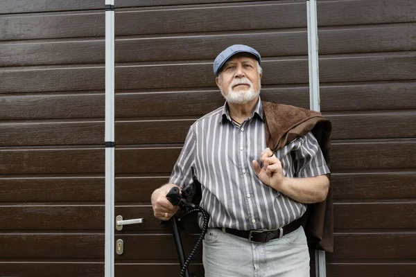 A positive, elderly man stands near the door and holds an eco scooter behind the wheel. Healthy lifestyle of the elderly.