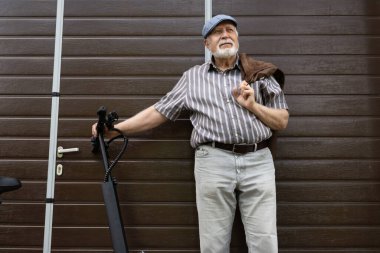 Olumlu, yaşlı bir adam kapının yanında duruyor ve direksiyonun arkasında bir eko scooter tutuyor. Yaşlıların sağlıklı yaşam tarzı.