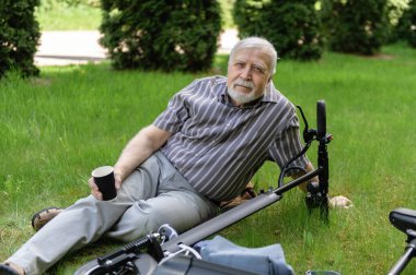 Olumlu, yaşlı bir adam parkta yürüyüşe çıktı, durdu ve elektrikli bir scooterın yanındaki çimlerin üzerinde dinlenmek için uzandı. Yaz, güneşli bir gün. Yaşlıların sağlıklı yaşam tarzı.