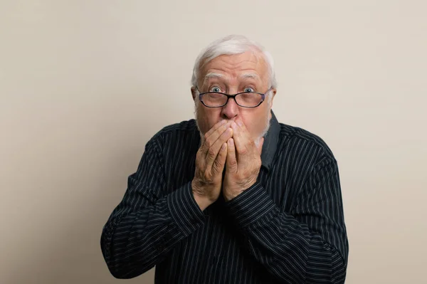 elderly gray-haired man with a beard in glasses and a black shirt covered his mouth with his hands, scared, emotions in the frame, waist portrait, light background, studio
