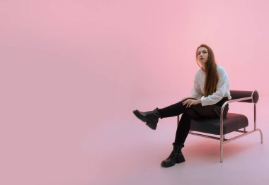 Çekici, uzun saçlı, sandalyede oturan ve çizme giyen genç bir kadının vücut ölçülerinde görüntüsü. Uzayı kopyala, pastel pembe arka plan. Ayakkabı mağazasına yönlendir. Dizinin bir parçası. Gelişigüzel.