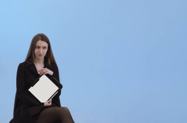 Siyah elbiseli, uzun saçlı genç bir kadın tabletinde beyaz ekran gösteriyor. Yeni uygulama ya da ürün tanıtımı. Kopya alanı olan açık mavi bir arkaplan. Dizinin bir parçası. Çevrimiçi eğitim, yakınlaştırma eğitimi.