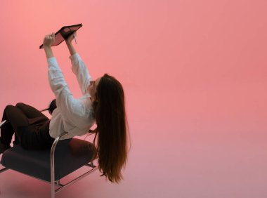 Sandalyede oturan genç bir kadın tabletini kaldırmış, düz elleriyle. Arkadan bak. Dikey, boşluğu kopyala. Pembe renkli arka planda izole edilmiş. Serbest tasarımcı ya da çizer. Bir serinin parçası.