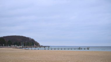 Geniş, boş kumlu sahil, uzak rıhtım ve orman burnu bulutlu gökyüzü altında, sakin alçak deniz, soluk renk paleti, minimal