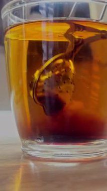 Çay demlenmiş cam bardak, kehribar girdabı ve çay poşeti ahşap masadaki sıcak suya renk salıyor.