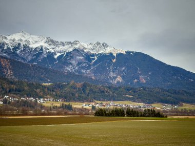 Karla kaplı tepeleri ve geniş tarım arazisi önü ile bulutlu Alp Vadisi Dağılmış Köy Evleri