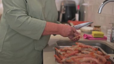 Kısa beyaz saçlı yaşlı kadın titizlikle mutfak makası ile taze çiğ karides hazırlıyor. Akşam yemeği için lavabonun yanında deniz ürünlerini temizliyor.